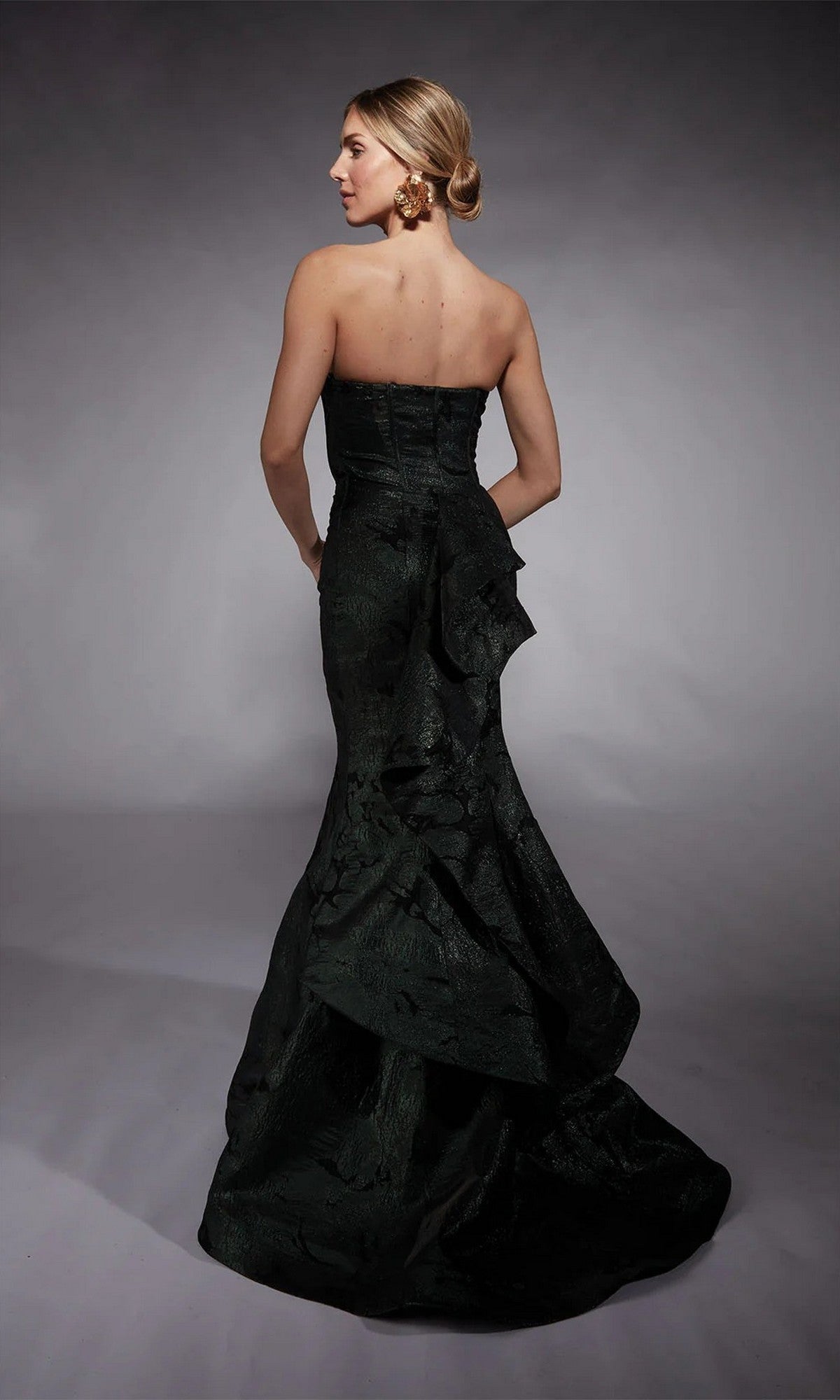 A woman models the Long Formal Dress: Alyce 27747, featuring a strapless corset bodice, dark textured mermaid skirt, and ruffled back detail as she stands facing away against a plain backdrop.